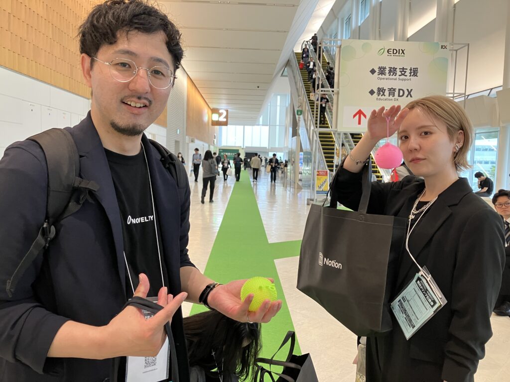 ノベルティ社員2名が、ブースでもらったピンクと黄色のふわふわの毛が生えたヨーヨーを持っている。