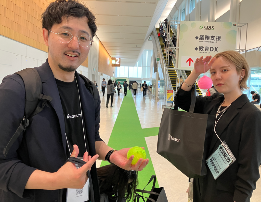ノベルティ社員2名が、ブースでもらったピンクと黄色のふわふわの毛が生えたヨーヨーを持っている。