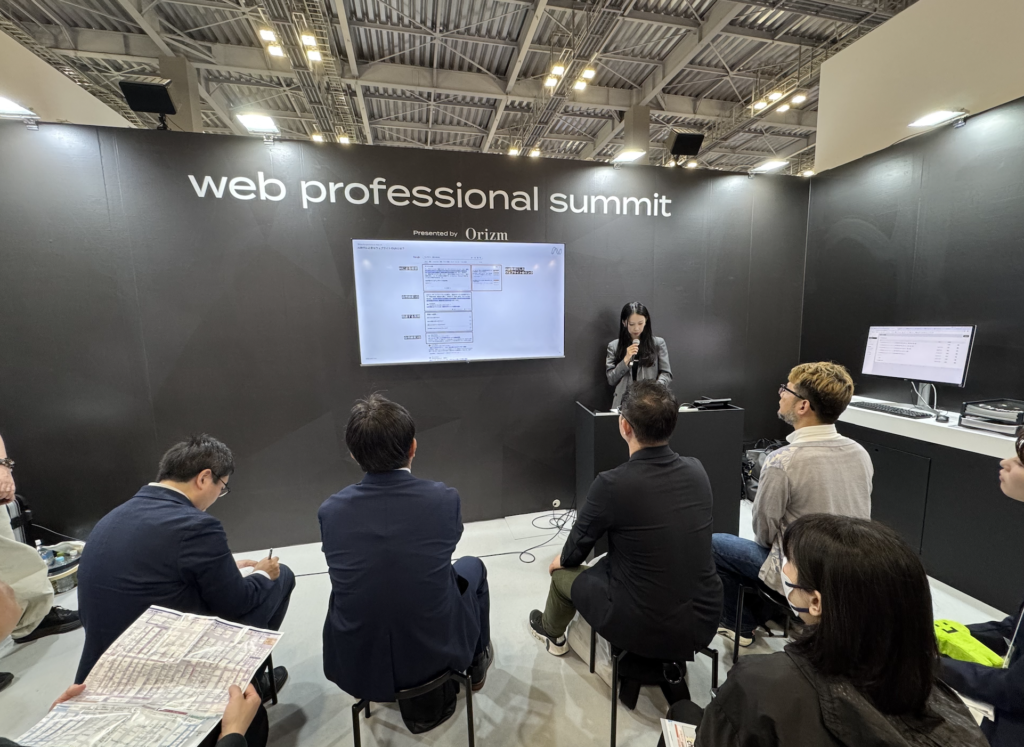 「web professional summit」と書かれたブース中央の登壇スペースにノベルティの社長がマイクを持って立っている。その前にセミナーを聞きにきた来場者が8名ほど、椅子に座ってセミナーを聞いている。
